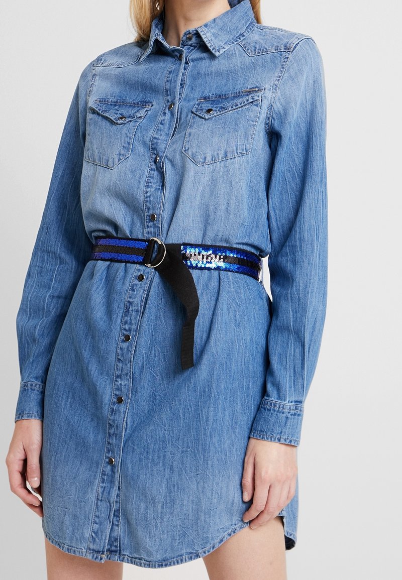 Femme portant une robe en jean bleu clair boutonnée avec des poches poitrine et une ceinture noire et bleue à sequins à la taille.