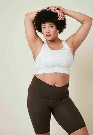 Femme aux cheveux bouclés en brassière de sport blanche et short foncé posant les bras levés sur un fond uni.