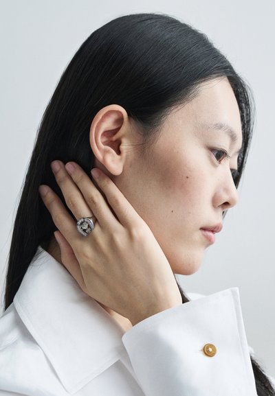 Ezüst gyűrű bonyolult mintázattal és díszítésekkel, egy kézen viselve, közel egy archoz, amelyen egyenes fekete haj van, fehér ingben öltözve.