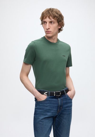 Jeune homme portant un t-shirt vert à manches courtes et un jean bleu avec une ceinture noire, debout les mains dans les poches devant un fond uni.