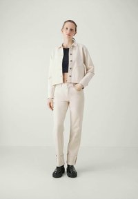 Giacca cropped beige chiaro con chiusura a bottoni, abbinata a pantaloni coordinati. L'orlo arrotondato, i polsini e le scarpe con piattaforma nera completano il look.