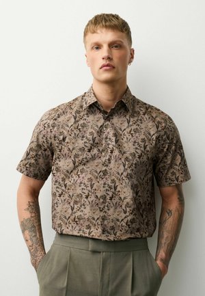 Mann mit kurzen blonden Haaren, der ein beige-blumiges Kurzarmhemd mit Knöpfen und graue Hosen trägt, Hände in den Taschen, vor einem einfarbigen Hintergrund.