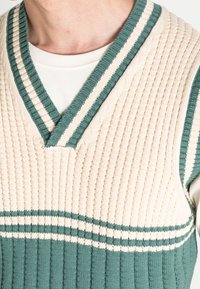 Ärmelloser Strickpullover in Creme und Petrol, mit V-Ausschnitt, gerippter Textur und horizontalen Streifen über der Brust und am Saum.