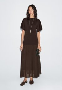 Braunes Zweiteiler-Outfit mit einem taillierten Top mit kurzen Ärmeln und einem fließenden, geschichteten Rock. Die Accessoires umfassen eine strukturierte schwarze Clutch und braune Schuhe.