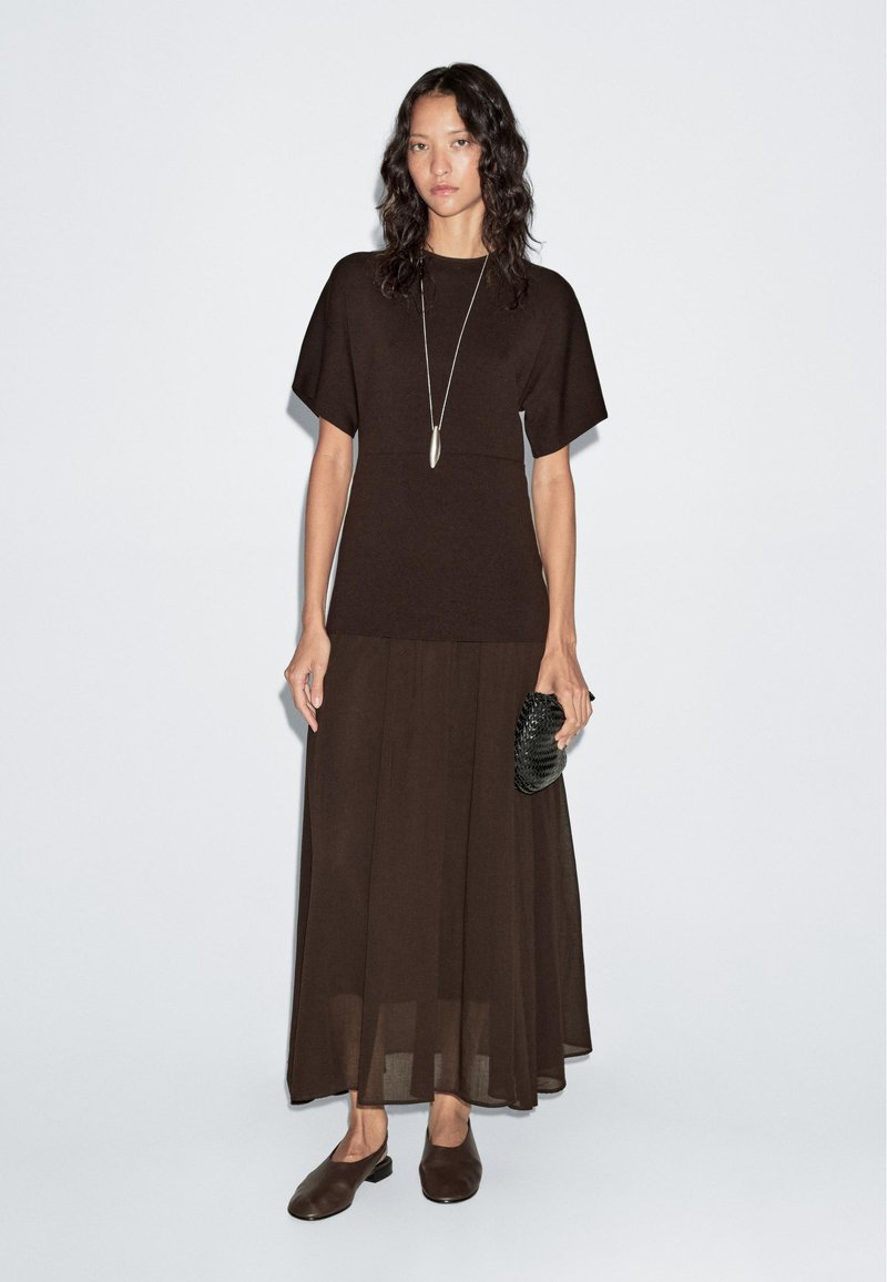 Braunes Zweiteiler-Outfit mit einem taillierten Top mit kurzen Ärmeln und einem fließenden, geschichteten Rock. Die Accessoires umfassen eine strukturierte schwarze Clutch und braune Schuhe.