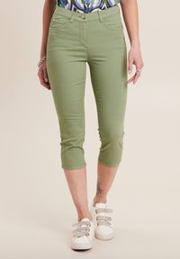 Pantaloni verdi cropped con un design aderente, realizzati in tessuto elasticizzato. Presentano cinque tasche e una chiusura con bottone frontale, abbinati a sneaker bianche.