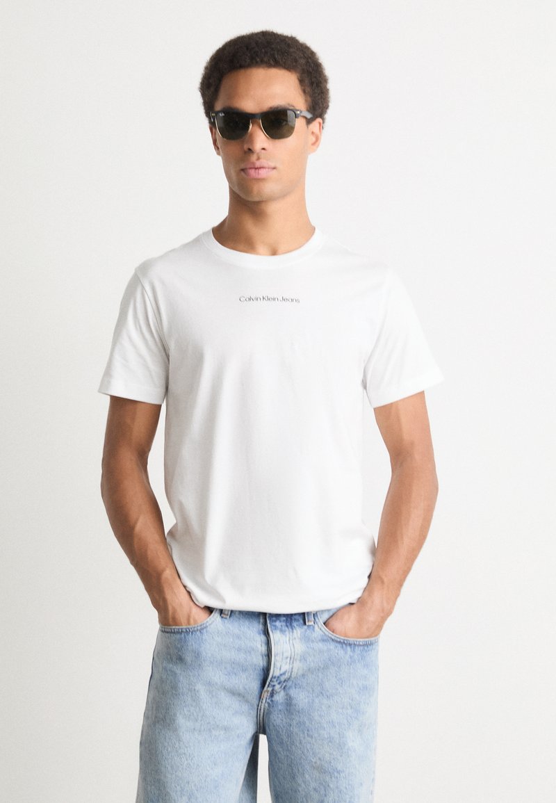 Bílý bavlněný tričko s centrálním logem Calvin Klein Jeans, v kombinaci s tmavými slunečními brýlemi a světle modrými džínami.