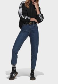 Černá zipová bunda s bílými pruhy, vysoko vytažené modré džíny, černé tenisky a pruhované ponožky. Kulatá spona opasku a zúžený design nohavic.