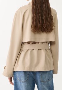 Manteau trench beige avec une taille ceinturée, des manches longues et un dos à plusieurs couches, associé à un pantalon en denim. Tissu lisse avec une texture minimale.