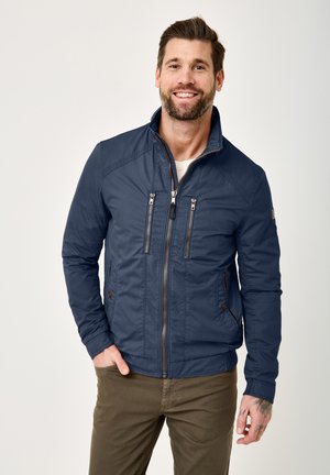 Mann lächelt, trägt eine marineblaue Jacke mit Reißverschluss mit zwei Brusttaschen und zwei Seitentaschen sowie olivgrüne Hose, steht vor einfachem hellem Hintergrund.