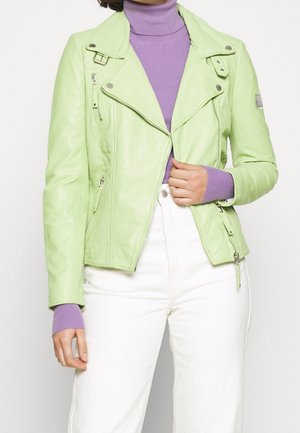 Woman wearing light green leather jacket over purple turtleneck sweater and white high-waisted pants, adjusting jacket zipper.