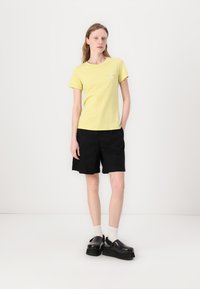 T-shirt en coton jaune à manches courtes, shorts noirs et chaussures noires massives. Les chaussettes sont blanches. Design simple, sans motifs ni éléments métalliques visibles.