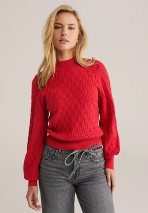 Pull en tricot rouge avec un motif de feuilles texturé, col et poignets côtelés. Associe à un pantalon gris avec un détail de taille nouée.