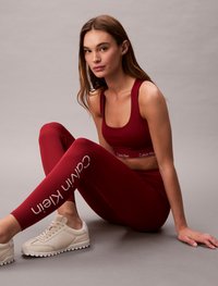 Bordeaux sports-bh og leggings med Calvin Klein-logo, kombinert med lysebeige sneakers, mot en nøytral bakgrunn.