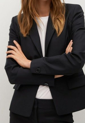 Frau mit verschränkten Armen, die einen schwarzen Blazer über einem weißen Oberteil trägt und eine professionelle und selbstbewusste Haltung vor einem einfarbigen Hintergrund zeigt.