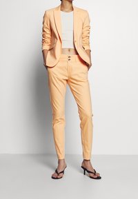 Peach-colored tailored suit featuring a fitted blazer and slim trousers. Textured fabric with two-button detail on the waistband. Worn with black sandals.