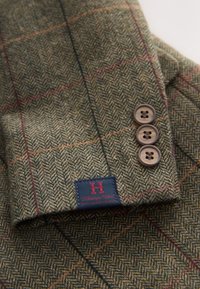 Brun fiskebensmønstret tweedjakkeærme med tre brune knapper og en navyblå label med teksten Heritage Club Finest Quality Apparel.