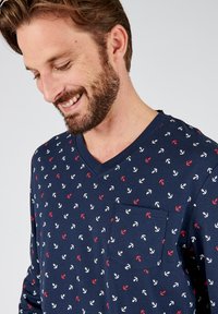 Maglietta a maniche lunghe blu navy con scollo a V, caratterizzata da un motivo ripetuto di ancore in rosso e bianco. Include una tasca frontale sul lato sinistro.