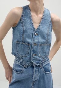 Jeanväst i ljusblått med djup V-hals, fem knappar och två framfickor. Har en kortad passform och synliga sömnadsdetaljer.