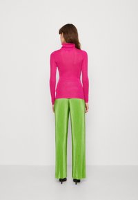 Žena stojící zády, oblečená v jasně růžovém dlouhém roláku a širokých limetkově zelených kalhotách s černými podpatky.