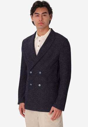 Uomo che indossa una giacca doppiopetto blu scuro sopra una camicia bianca abbottonata e pantaloni beige, in piedi davanti a uno sfondo bianco semplice.