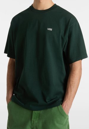 Mężczyzna ubrany w ciemnozielony t-shirt Vans z krótkim rękawem i jasnozielone spodnie, ręce w kieszeniach, na jednolitym tle.