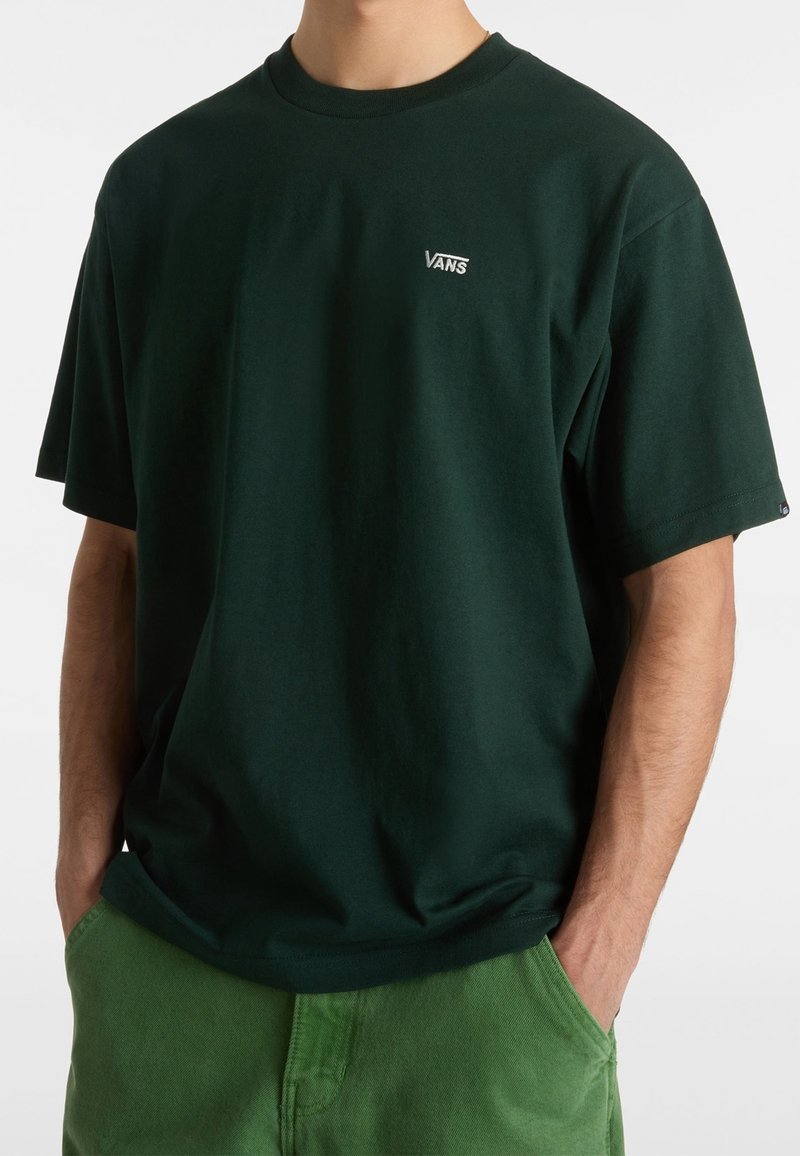 Homme portant un t-shirt Vans vert foncé à manches courtes et un pantalon vert clair, mains dans les poches, sur un fond uni.