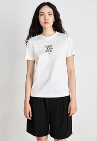 Tricou din bumbac alb, cu un logo negru care spune "Calvin Klein Jeans NYC." Tricoul are guler rotund și mâneci scurte.