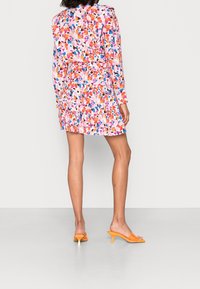 Robe à motif floral en rose, orange et bleu, avec des manches longues, un ourlet volanté et une silhouette ajustée. Le modèle porte des mules à bout ouvert orange.