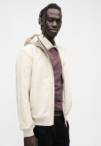 Bēša kapuce ar gludu apdari, priekšā ar rāvējslēdzēju un ribotām aprocēm, uzvilkta pāri tumši rūsganas krāsas džemperim.