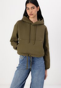 Ελιά πράσινη κοντή φόρμα με κορδόνι, τσέπη καγκουρό και κεντητές λεπτομέρειες. Συμπληρώνεται με ανοιχτό μπλε τζιν ίσια γραμμή.