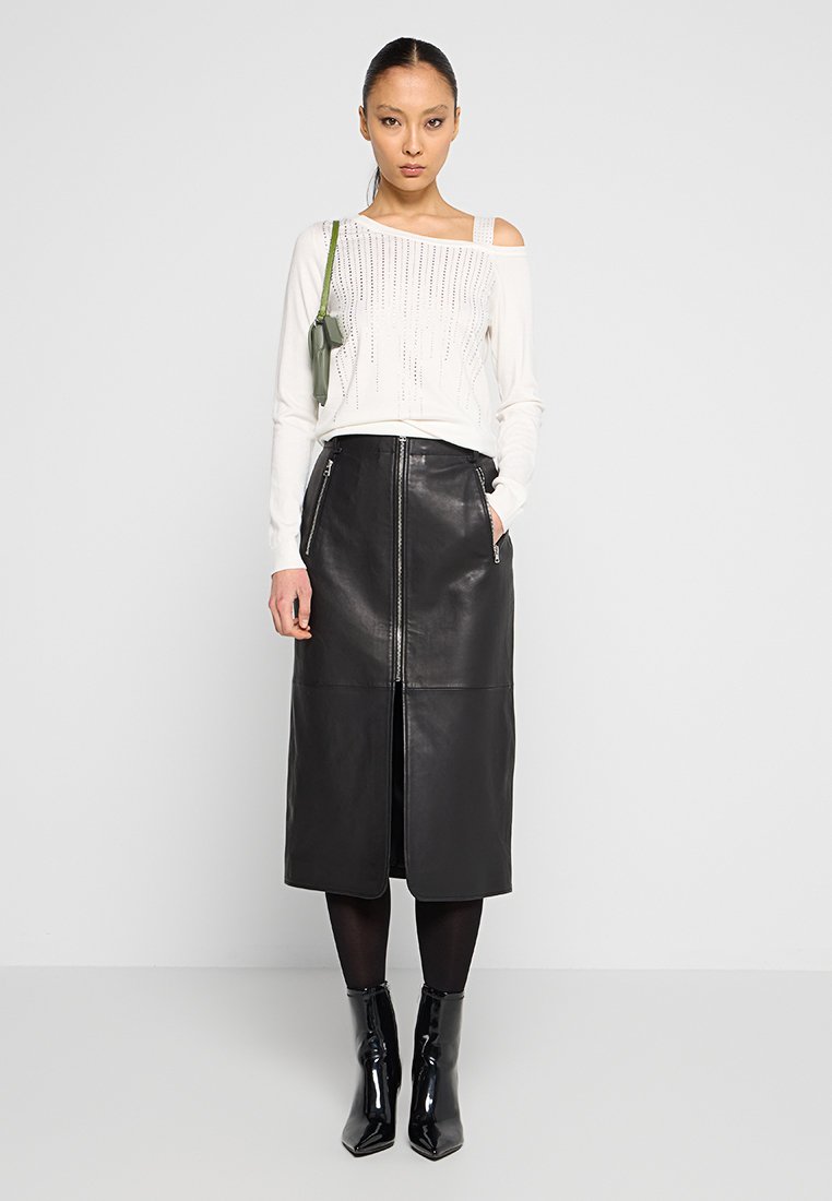 Weißer Off-Shoulder-Pullover mit einem bestickten Muster, kombiniert mit einem schwarzen Leder-Midirock mit einem Frontreißverschluss und Taschen. Schwarze Stiefeletten.