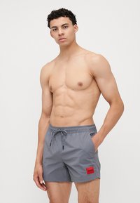 Shorts de bain gris avec une taille élastique, un cordon de serrage, et une étiquette rectangulaire rouge arborant le logo "HUGO". Le matériau semble léger et lisse.