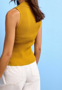 Mosterdgele mouwloze geribbelde top met een kraag, aansluitend ontwerp, gecombineerd met witte high-waisted broeken, tegen een blauwe achtergrond.