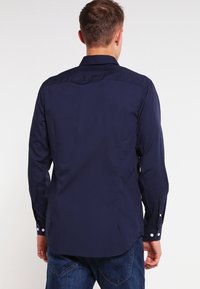 Tmavě modrá košile s dlouhým rukávem, vybavená límečkem na knoflíky, zadní částí s klínem a nastavitelnými manžetami, vyrobená z hladké látky s přizpůsobeným střihem.