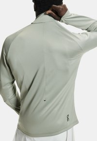 Langarm-Sportshirt in Hellgrün mit Stehkragen, glatter Textur und einem Logo unten links.