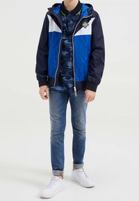 Sininen ja laivansininen tuulenpitävä takki, jossa valkoiset yksityiskohdat, vetoketju ja huppu, päällä tumman camo-t-paidan kanssa. Kapea malli siniset farkut ja valkoiset lenkkarit.