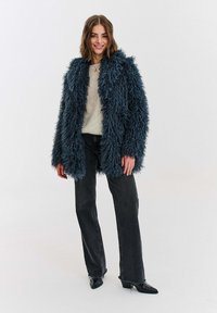 Žena v huňatom tmavomodrom kabáte, krémovom svetri, čiernych džínsoch a čiernych špicatých topánkach stojaca pred jednoduchým bielym pozadím.