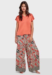 Korallefarbene Satin-Bluse mit kurzen Ärmeln, kombiniert mit weit geschnittenen Hosen, die rot-schwarze Blumenmuster und einen schwarzen gemusterten Saum aufweisen.