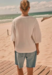 Person with hair in a bun wearing a white loose shirt and denim shorts standing on a wooden path facing a sandy beach and ocean.