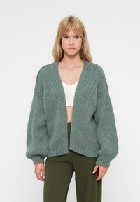 Cardigan lavorato a maglia di un verde chiaro con una trama spessa, design aperto davanti e maniche voluminose. Indossato sopra un top bianco e pantaloni verdi.