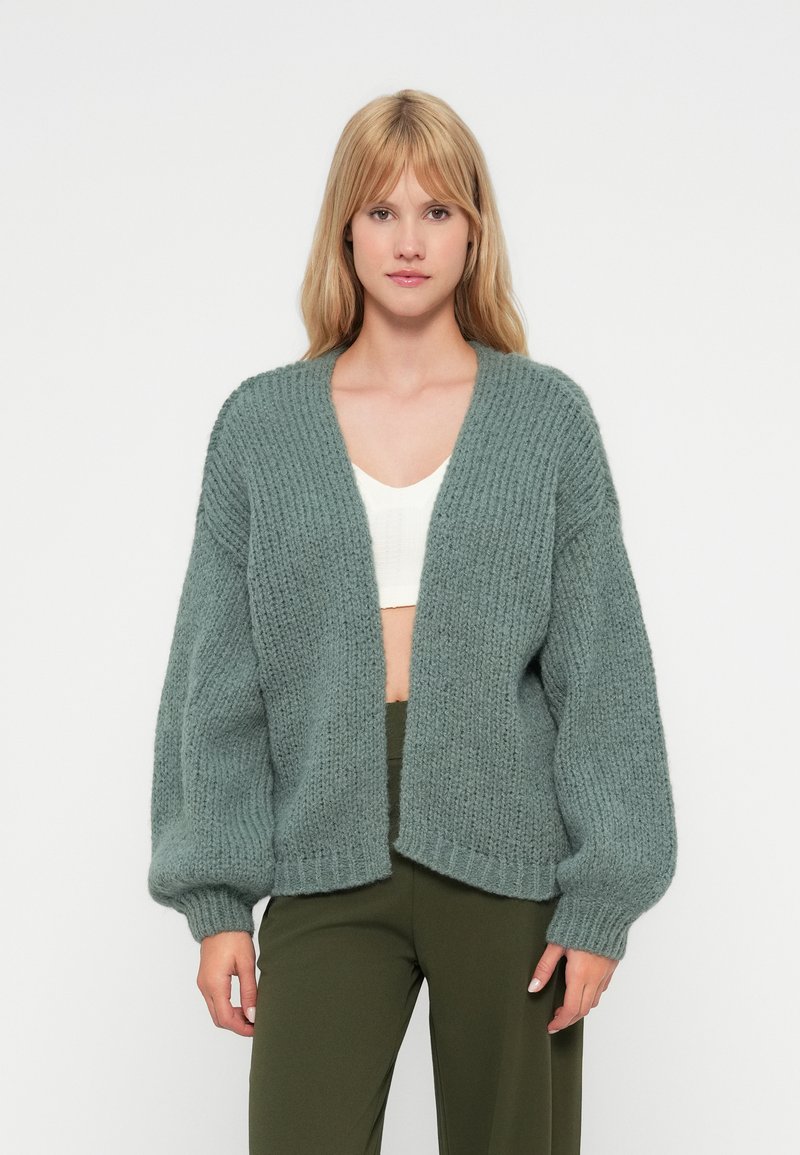 Cardigan lavorato a maglia di un verde chiaro con una trama spessa, design aperto davanti e maniche voluminose. Indossato sopra un top bianco e pantaloni verdi.