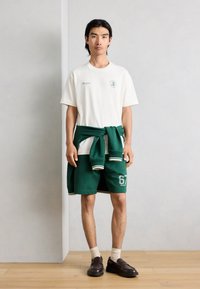 Νέος άνδρας που στέκεται μέσα σε κλειστό χώρο, φορώντας λευκό μπλουζάκι, πράσινα σορτς, πράσινο φούτερ δεμένο γύρω από τη μέση, μπεζ κάλτσες και σκουρόχρωμα καφέ παπούτσια.