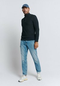 Donker gebreide trui met hoge kraag en drie knopen, gecombineerd met lichtblauwe jeans en witte sneakers. Man draagt een lichtblauwe pet.