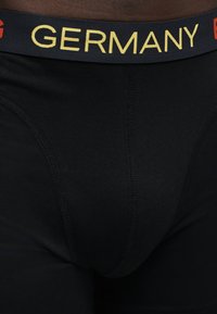 Μαύρα boxer briefs με μαύρη ζώνη μέσης που έχει τη λέξη "GERMANY" με κίτρινα γράμματα μπροστά.