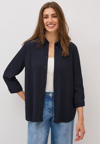 Mørkeblå skjorte med knaplukning i et tekstureret stof, med krave og ¾-ærmer, kombineret med lyseblå jeans.