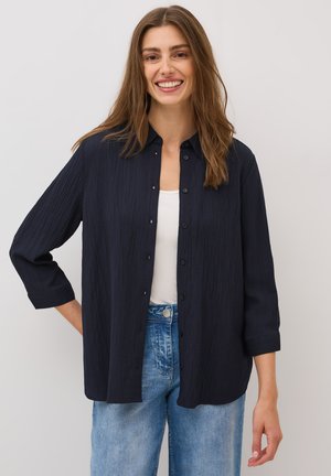 Chemise bleu marine à boutons avec un tissu texturé, comportant un col et des manches trois-quarts, associée à un jean bleu clair.