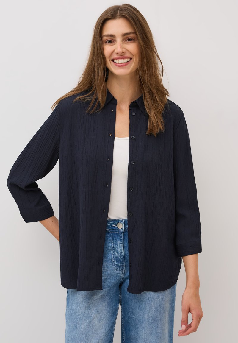 Mørkeblå skjorte med knaplukning i et tekstureret stof, med krave og ¾-ærmer, kombineret med lyseblå jeans.