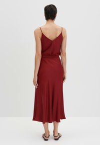 Bordeauxfarbendes Satin-Kleid mit Spaghettiträgern, V-Ausschnitt auf dem Rücken, tailliertem Design und einem ausgestellt geschnittenen Rock, der bis zur Mitte der Wade reicht. Das Modell trägt schwarze Sandalen.