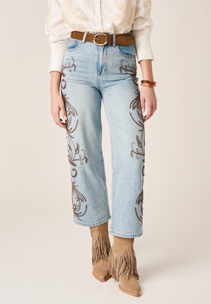 Jasnoniebieskie dżinsy z brązowym kwiatowym haftem na nogawkach, noszone z beżowymi zamszowymi botkami z frędzlami, brązowym paskiem, kremową bluzką i bursztynowymi bransoletkami.